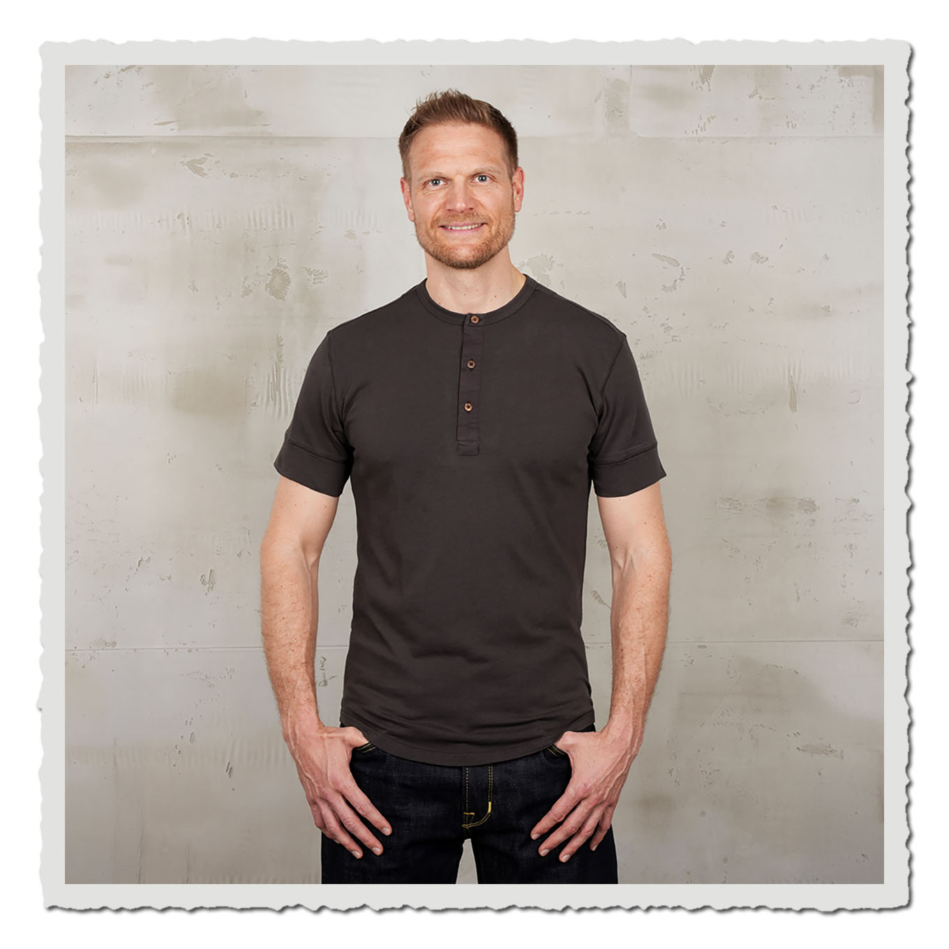Person mit dunkelbraunem Kurzarmshirt und Jeans vor hellem, neutralem Hintergrund im Studio.